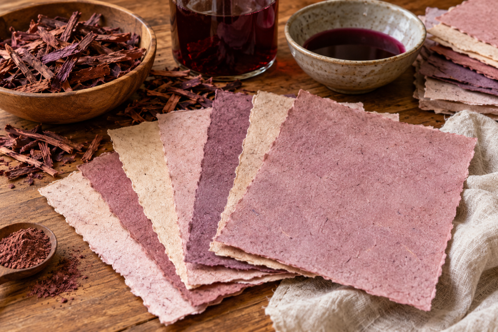 Handmade paper sheets in pink and earthy tones beside Mimosa Hostilis root bark and botanical dye materials.